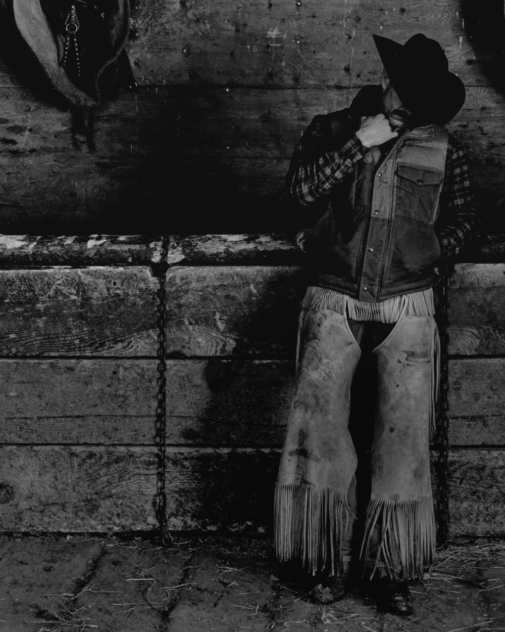 Black-and-white photograph of a cowboy in chaps standing against a wall